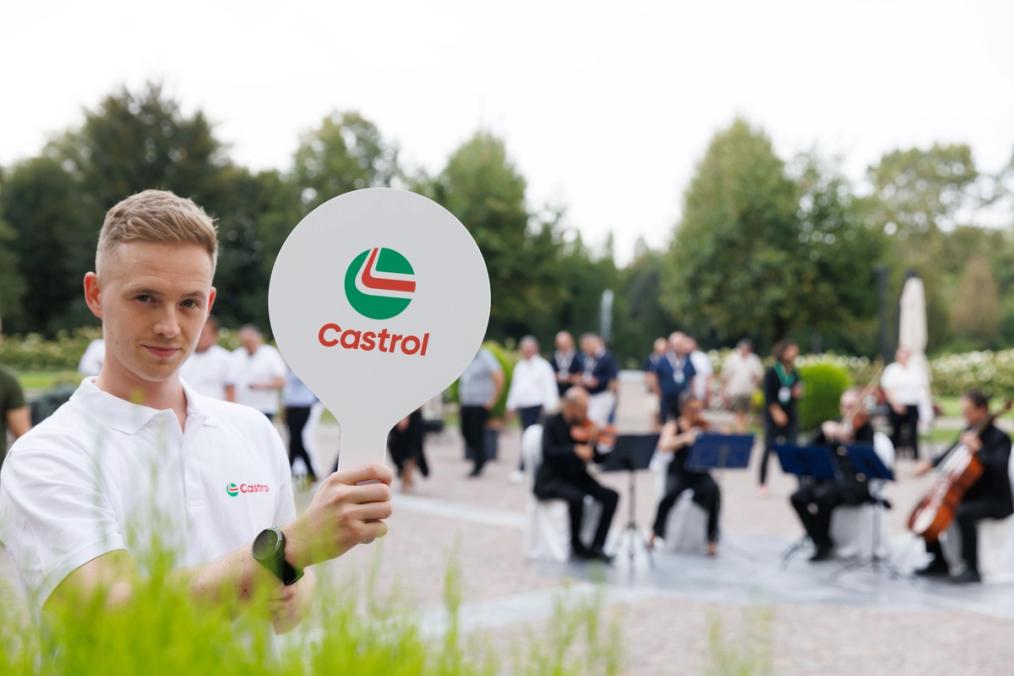 Mand i en hvid Castrol polo holder et Castrol-skilt op udenfor, mens der spilles klassisk live musik i baggrunden