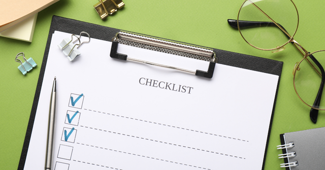 Overhead view of a checklist on a clipboard with a pen, glasses, binder clips, and a notebook on a green background.