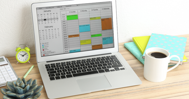 Laptop displaying a color-coded weekly calendar next to a coffee mug, alarm clock, calculator, and notebooks on a desk.