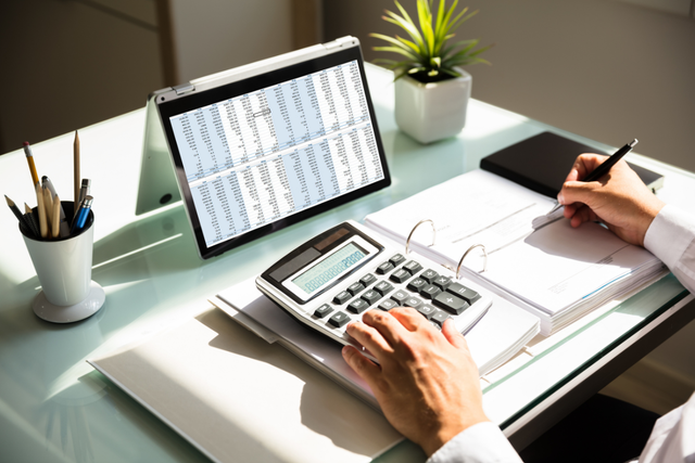 Person using a calculator and writing in a ledger at a glass desk, with a laptop showing a spreadsheet, a cup of pencils, glasses, and a potted plant.