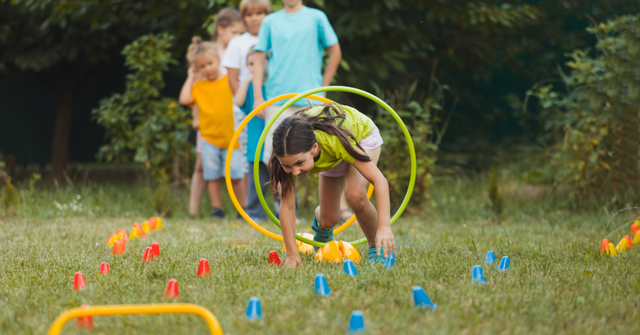 A young girl crawling through a hoop during an outdoor obstacle course, with other kids waiting in line behind her on the grass