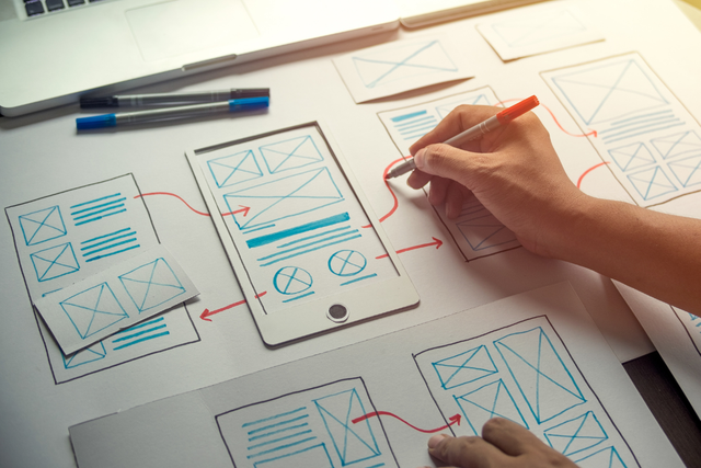 Person sketching mobile app wireframes connected by red arrows on paper, mapping out a screen flow with pens and a laptop on the desk.