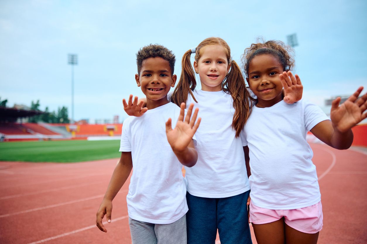 Happy little friends embracing waving during PE at the stadium and looking at camera.