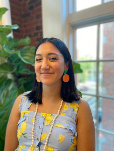 <A tan woman with short black hair and orange earrings smiles in front of window.>