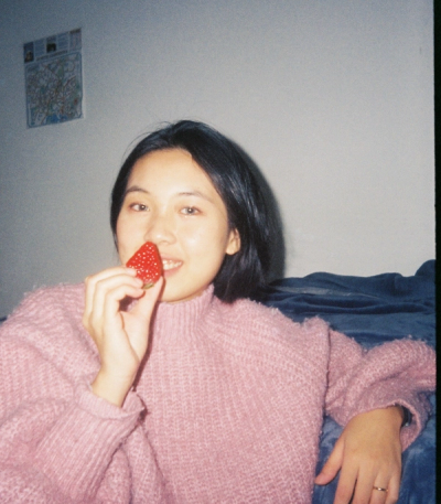 <A woman with black hair in a pink sweater holding a strawberry up to her nose and smiling.>