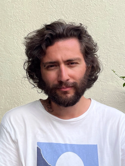 <A person in white shirt with long black hair and beard smiles softly in front of a plain tan background.>>