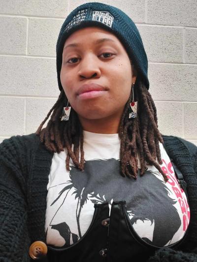 <A woman with mid length braids wearing a beanie. She is standing in front of a plain looking cinderblock wall.>