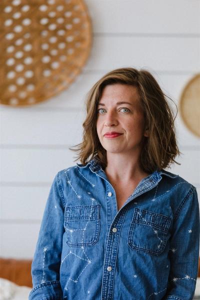 <A person with medium-length brown hair wearing a denim shirt looks at the camera with a white wooden background.>