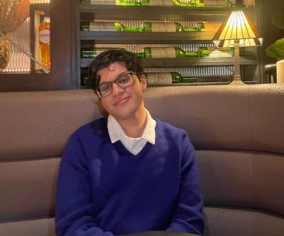 <A young man with glasses and dark hair smiles while sitting in a cushioned booth seat.>