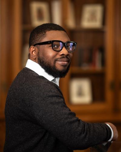 <A man with glasses and short hair in a sweater and collard shirt sitting in front of a wooden bookshelf.>