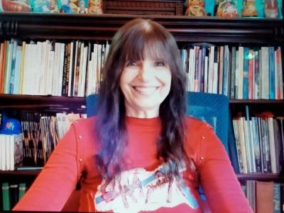 <A woman in a red sweater with long dark hair sits smiling at her desk with bookshelves in the background.>