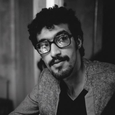 <A man with short curly dark hair, glasses, and a short beard looks past the camera in a black and white photograph.>