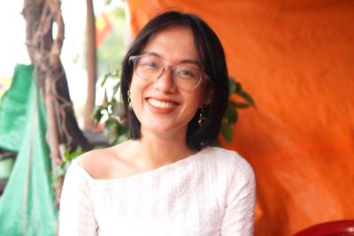 <A woman with glasses and short, dark hair stands smiling in front of an orange wall.>
