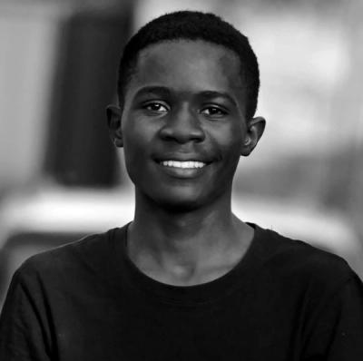 <A black and white photo of a man with short, dark hair standing in front of a blurring background and smiling.>