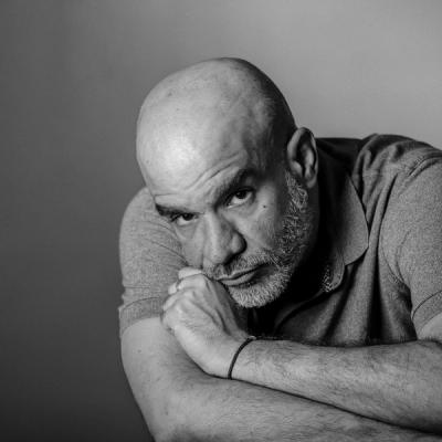 <A black and white photo of a bald man with a short grey beard crossing his arms and leaning forward onto a table.>