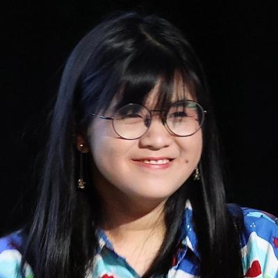 <A person with long, straight, dark hair, and glasses stands smiling in front of a black background.>