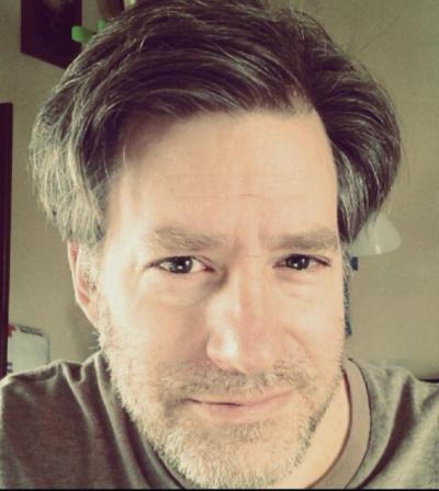 <A man with brown hair and short beard smirks at the camera with an office in the background.>