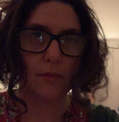 <A woman with glasses and short dark hair looking towards the camera in a dimly lit room.>