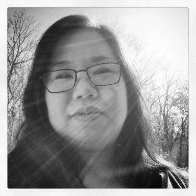 <A woman with shoulder-length black hair and glasses looks down as the sun shines above her.>