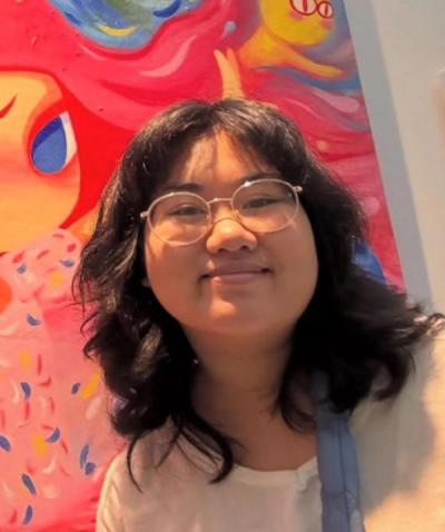 <A person with shoulder-length dark hair and glasses smiles with brightly colored artwork in the background.>