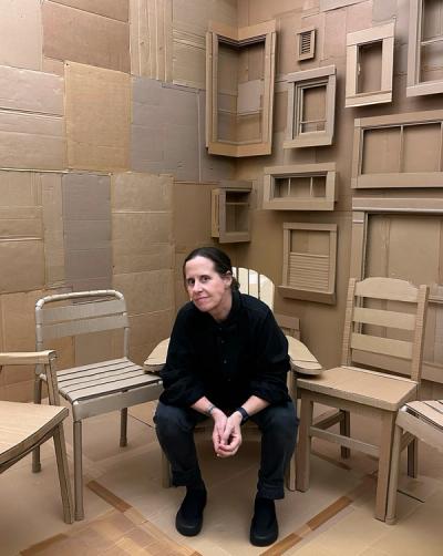 <A woman with dark hair sits in a chair in a room seemingly made entirely of cardboard.>