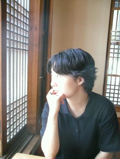 <A person with mid length black hair sits at table looking out the window in bright light.>