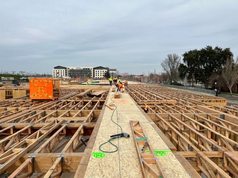 Workers installing open-web floor trusses with OSB sheathing on a large commercial building rooftop with Sacramento area buildings in the background by Mike Walker Lumber Co Inc