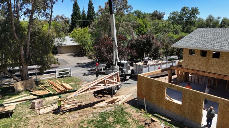 Boom truck setting residential roof trusses on a new home addition with existing roofline visible and workers guiding the truss into position in Sacramento by Mike Walker Lumber Co Inc