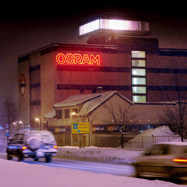 A snowy night scene featuring a building with a glowing red "OSRAM" sign, a small snow-covered house, and blurred cars on the road.