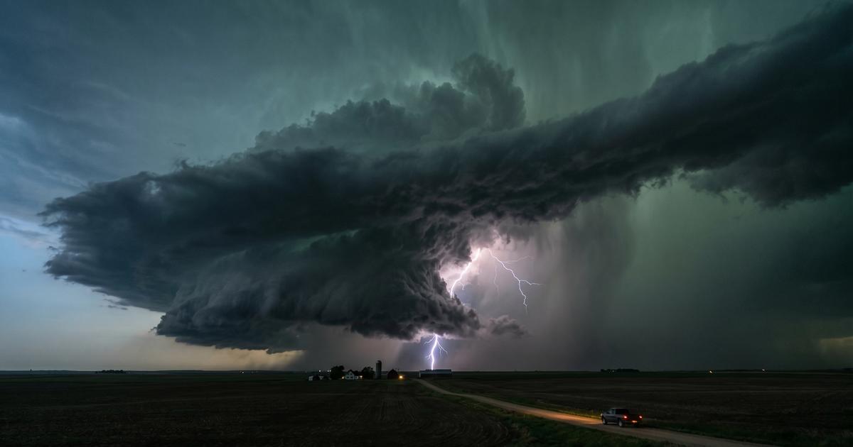 A dramatic cinematic wide shot of a massive dark supercell thunderstorm towering over a flat rural landscape at dusk, lightning illuminating the enormous rotating cloud formation from within, green-tinted sky with ominous shelf clouds extending to the horizon, severe weather atmosphere, cinematic editorial style, 16:9 aspect ratio, photorealistic, no text visible, no real logos, no real faces