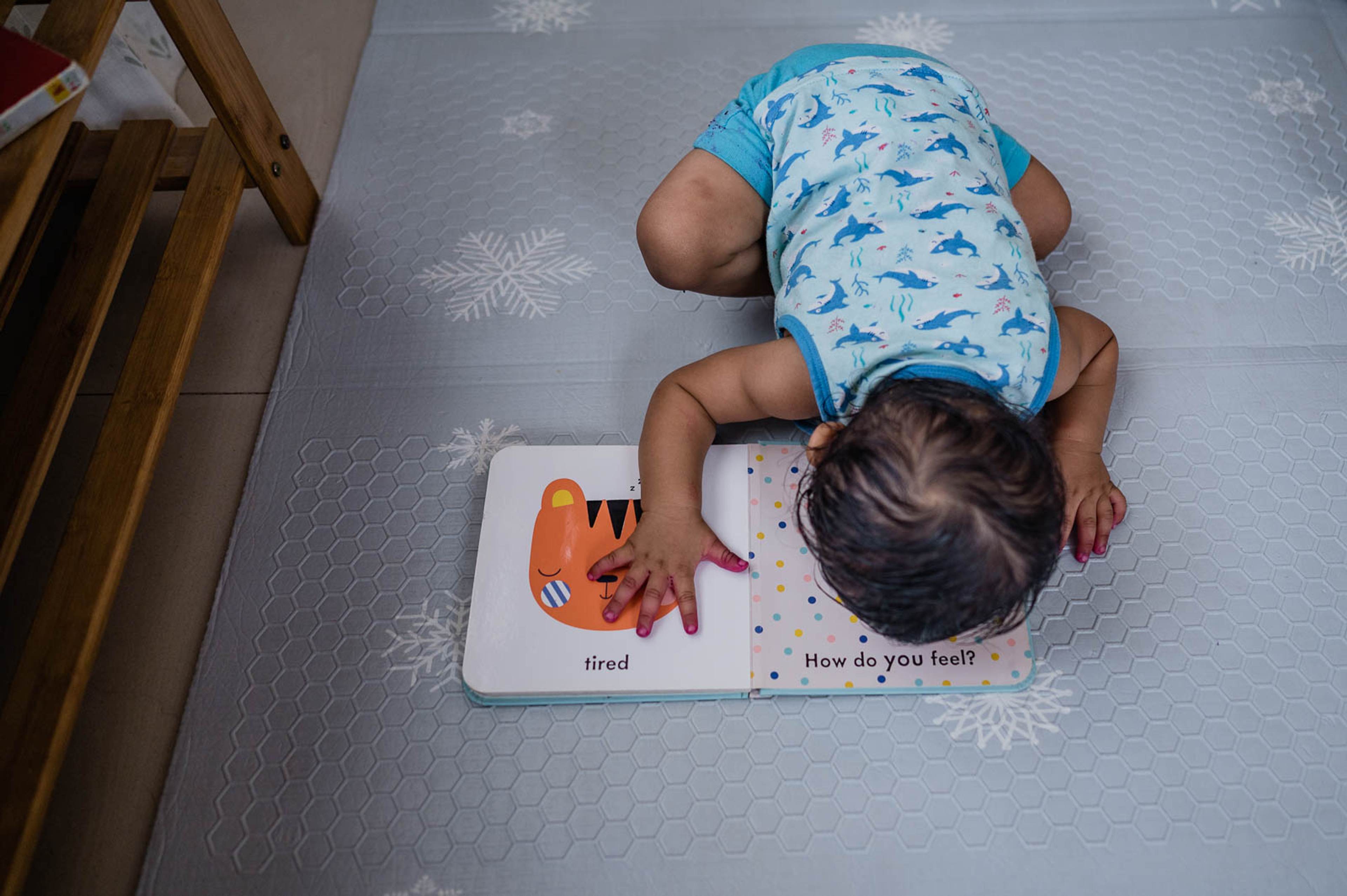Toddler in blue shirt lying on floor reading a children's board book, overhead view