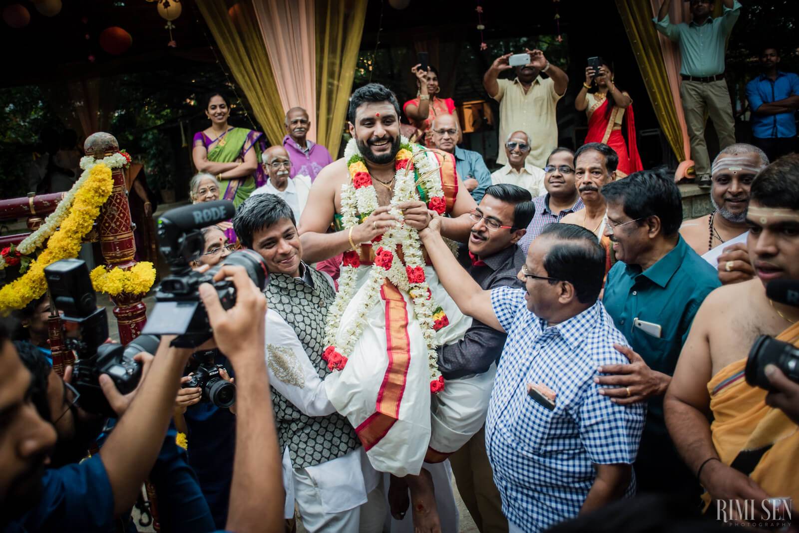 Groom in floral garland surrounded by joyful guests and photographers at Bangalore wedding celebration