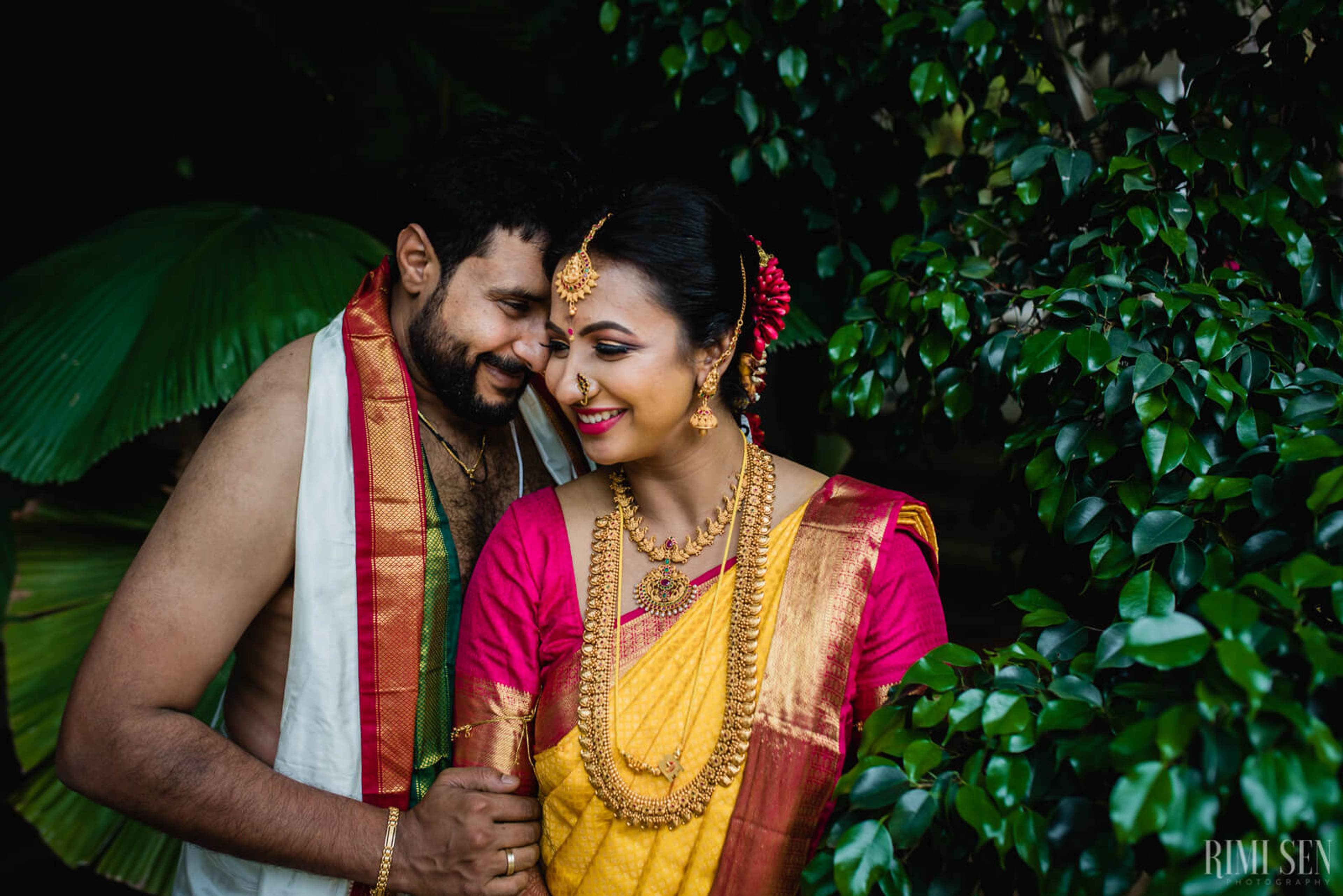 Nivetha and Sridhar in traditional South Indian attire, smiling together amid lush greenery in Bangalore, by Rimi Sen