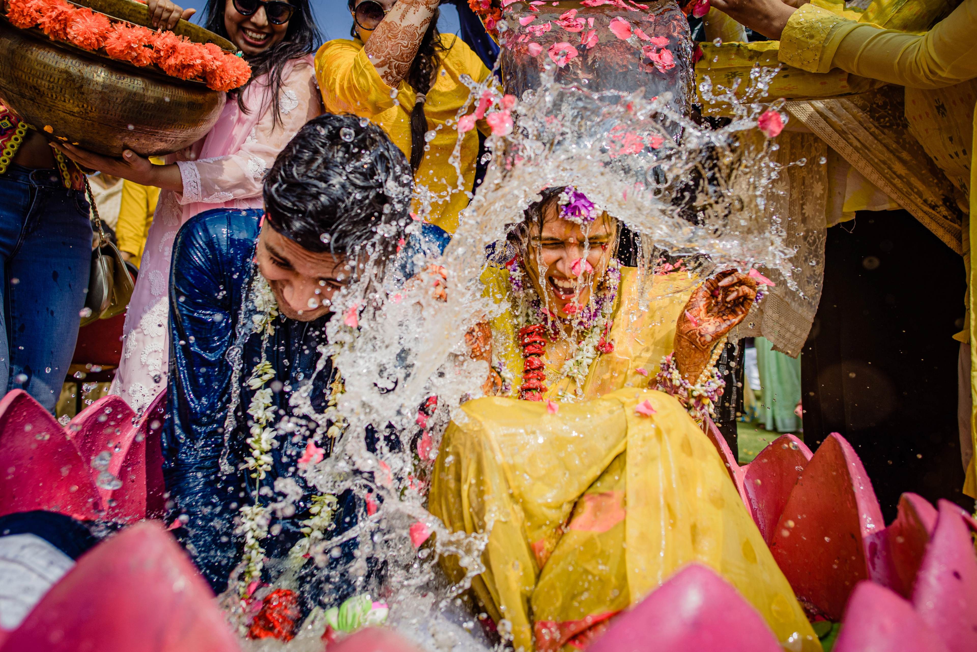 Couple in bright yellow and blue attire splashed with colorful flower petals and water during celebration