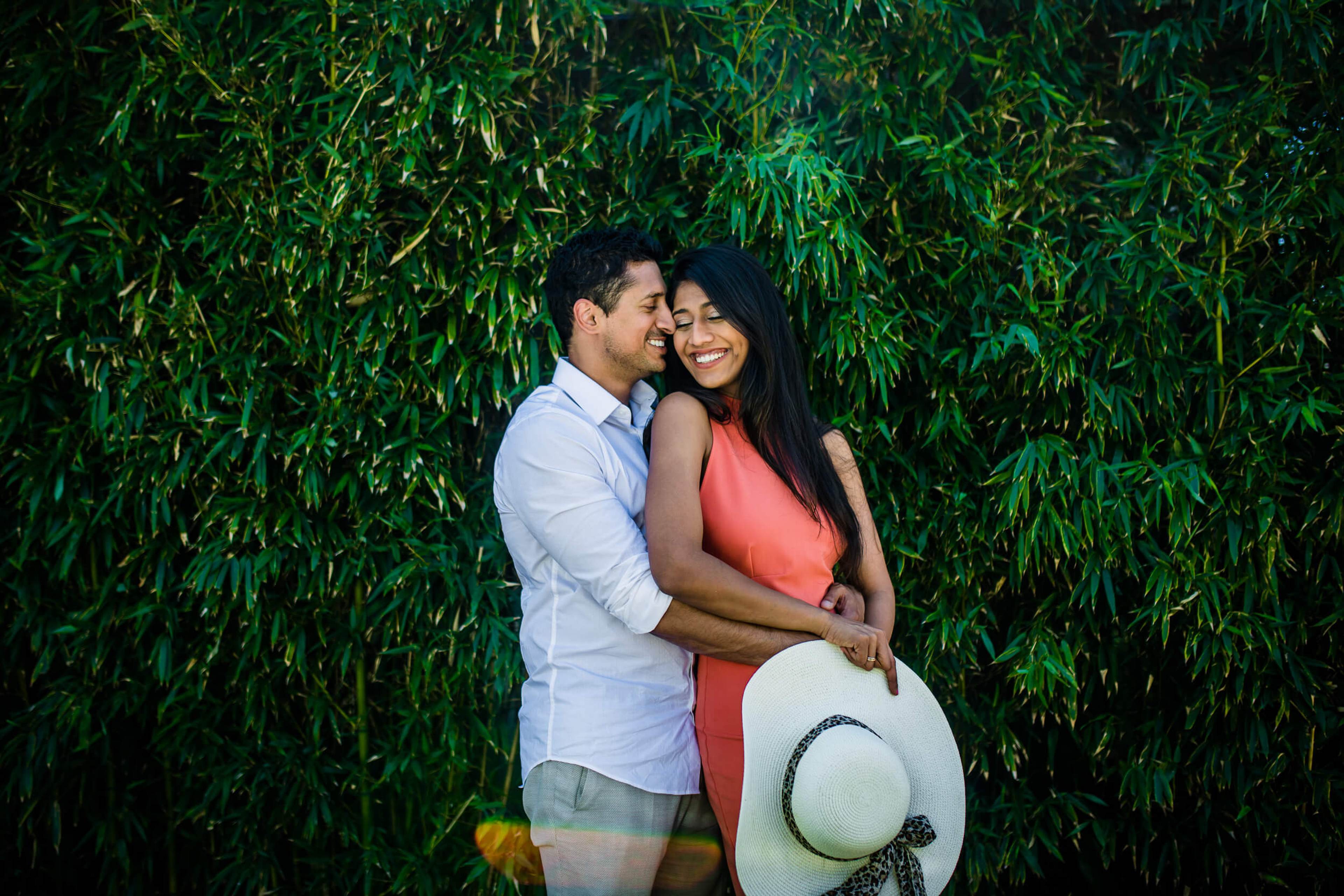 Anu and Raghu embrace against lush green foliage in California, holding a wide-brimmed hat