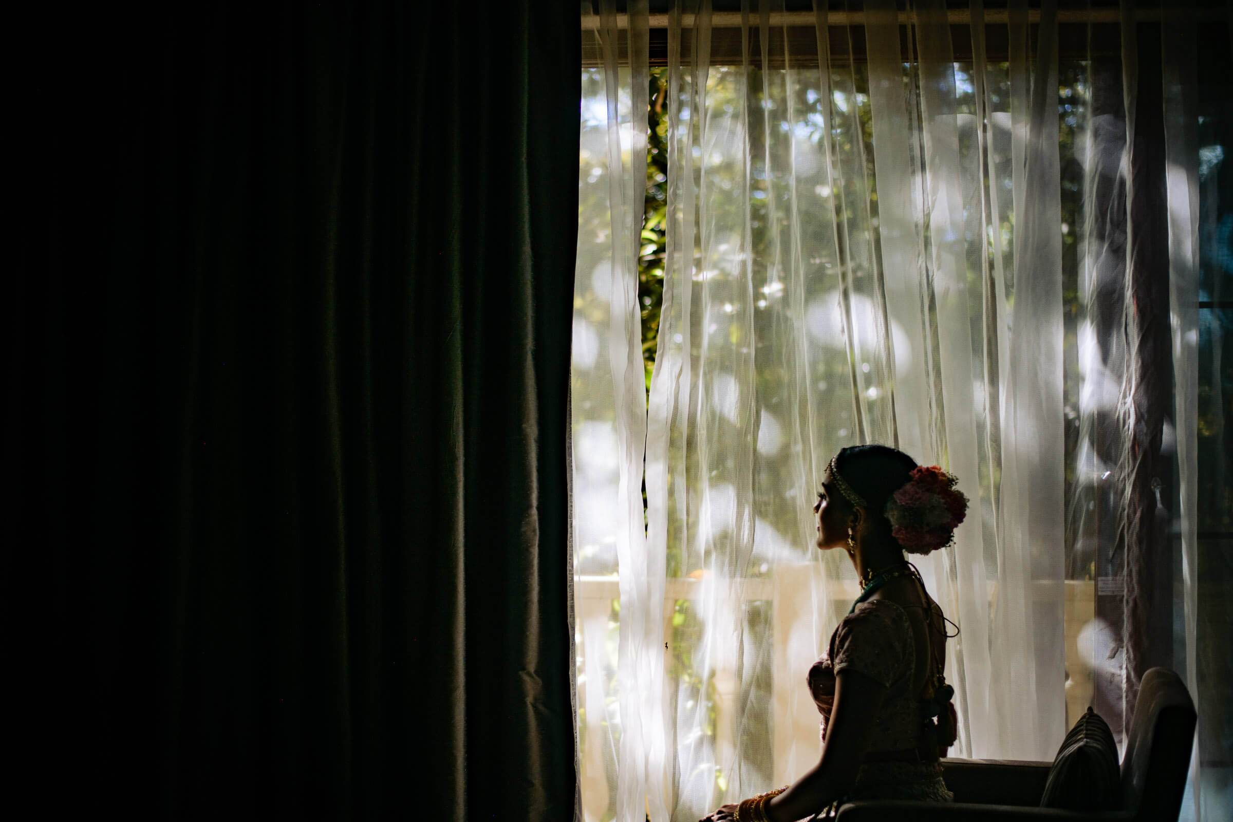 Shreya silhouetted at a bright window with sheer curtains and greenery visible behind