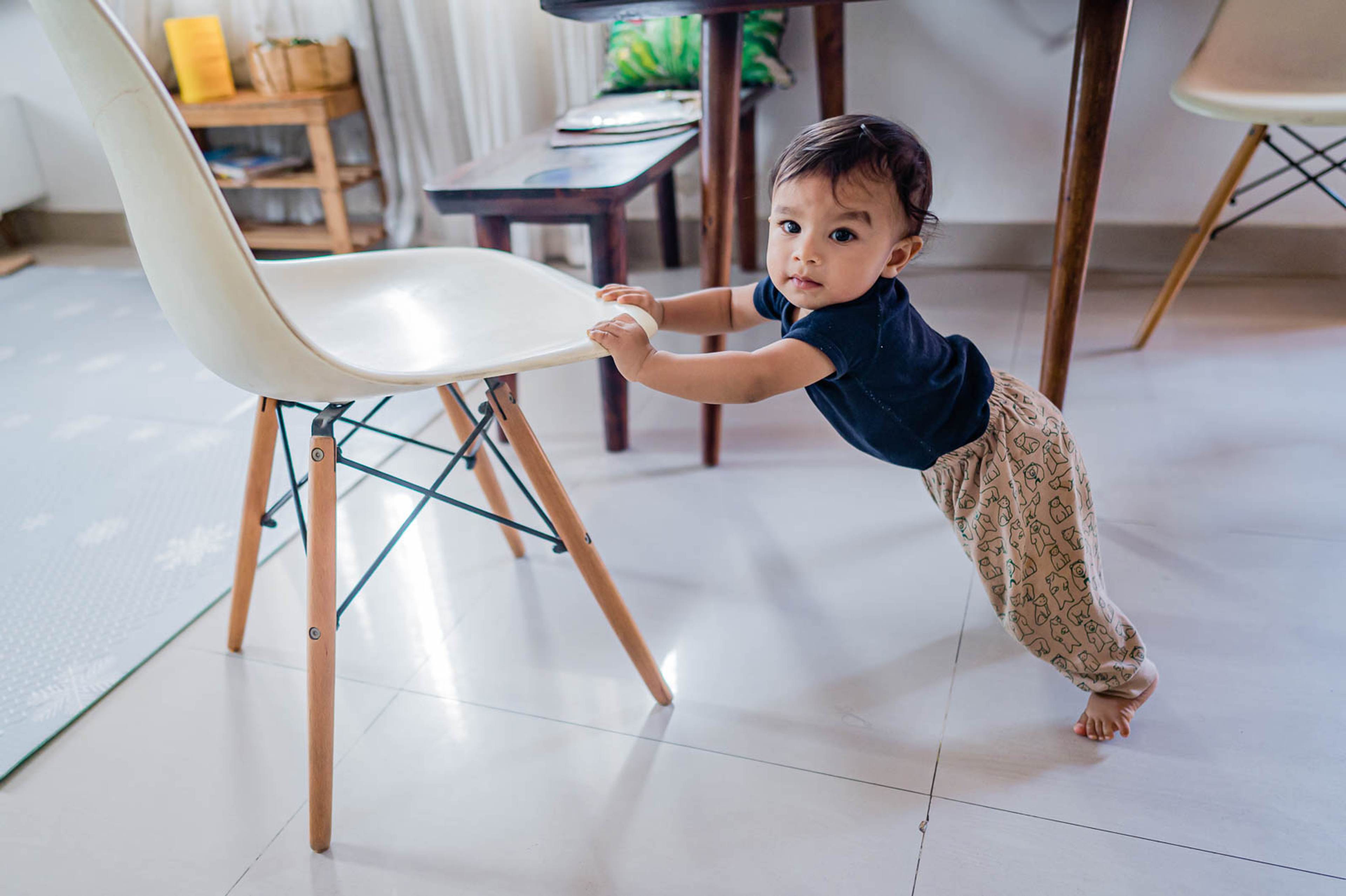 Toddler in dark shirt and beige pants reaching toward modern white chair in bright home interior