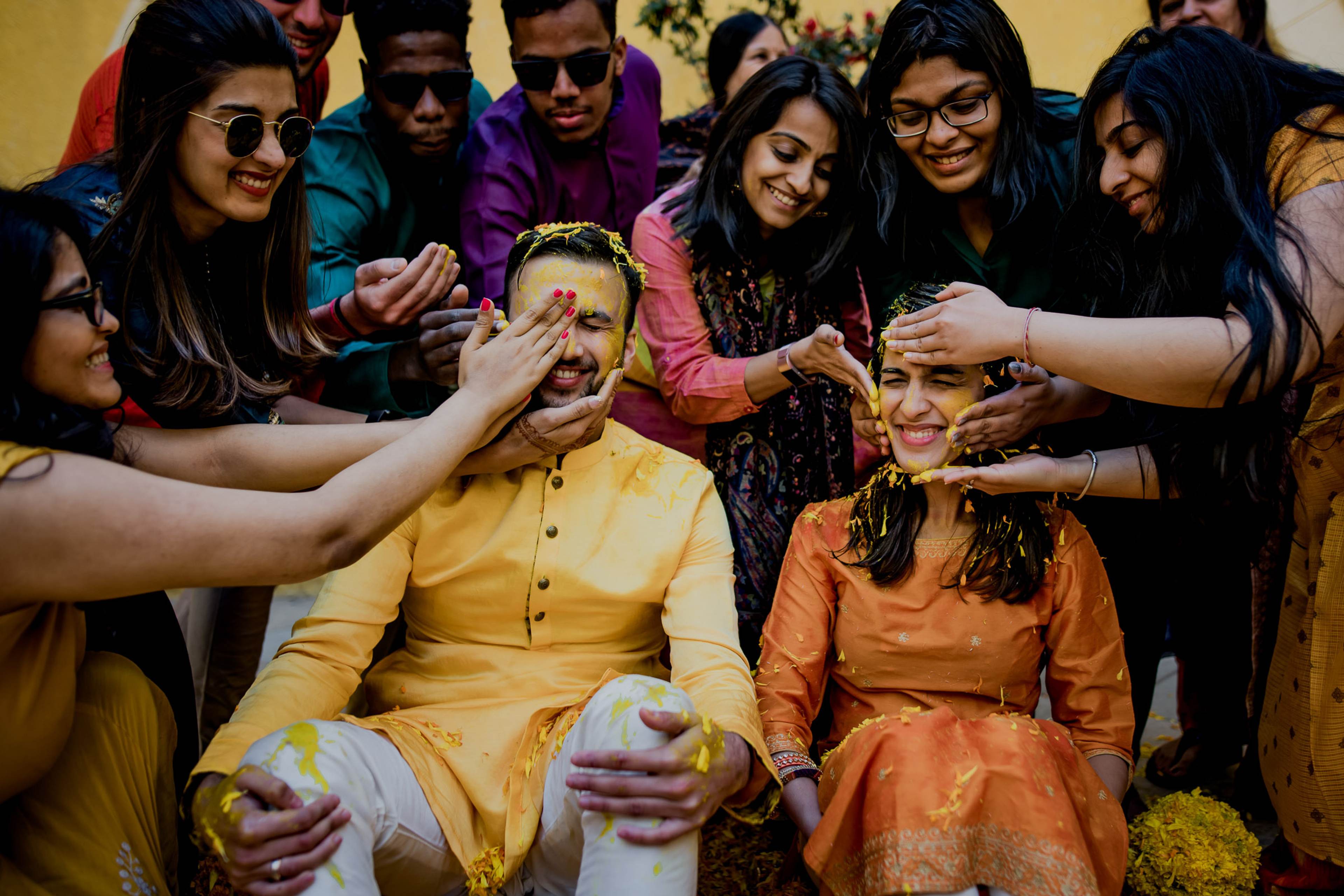 Group of friends playfully applying colored powder to couple seated in yellow and orange traditional attire during Holi celebration