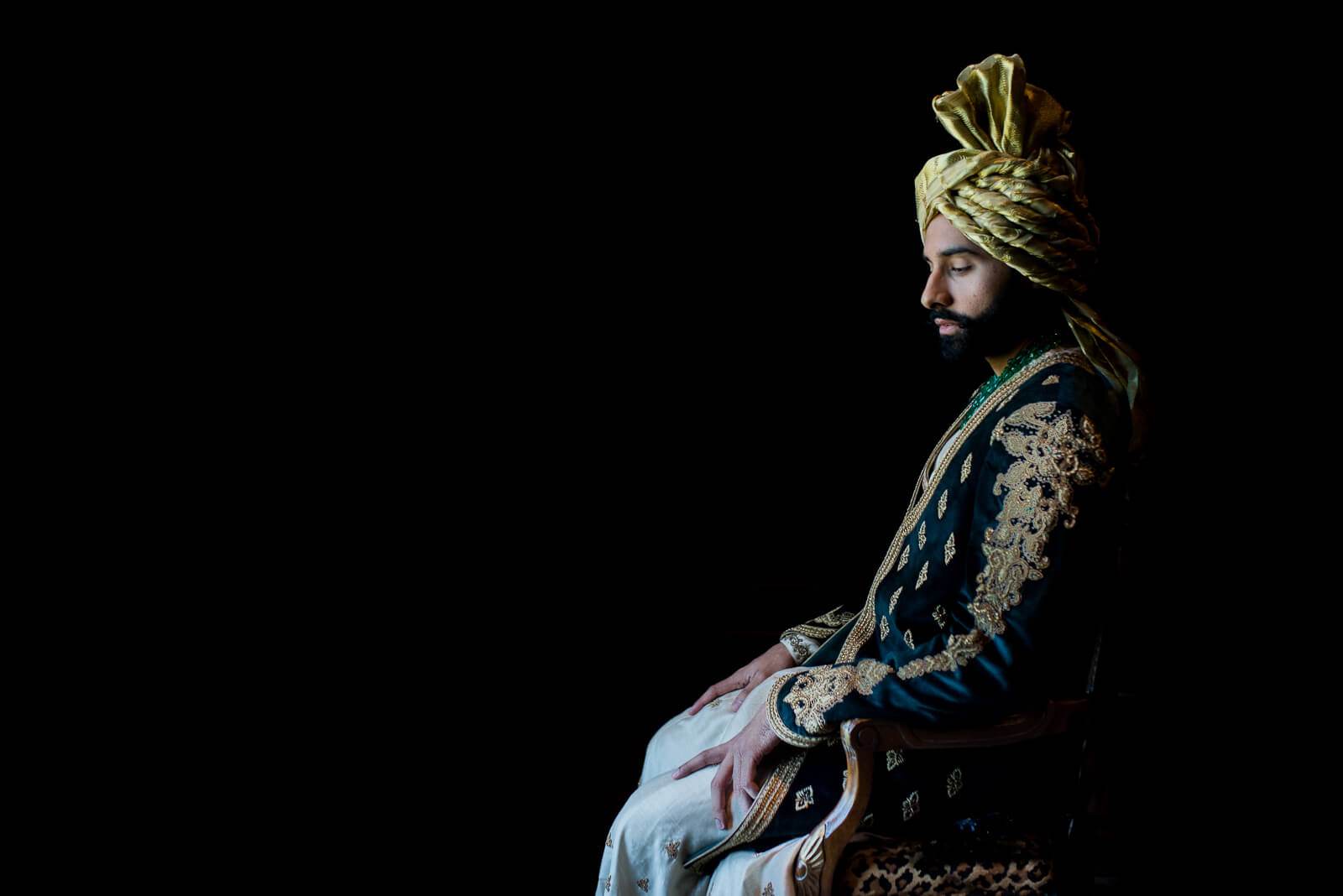 Ankit in ornate gold turban and embroidered navy jacket, seated profile against black background