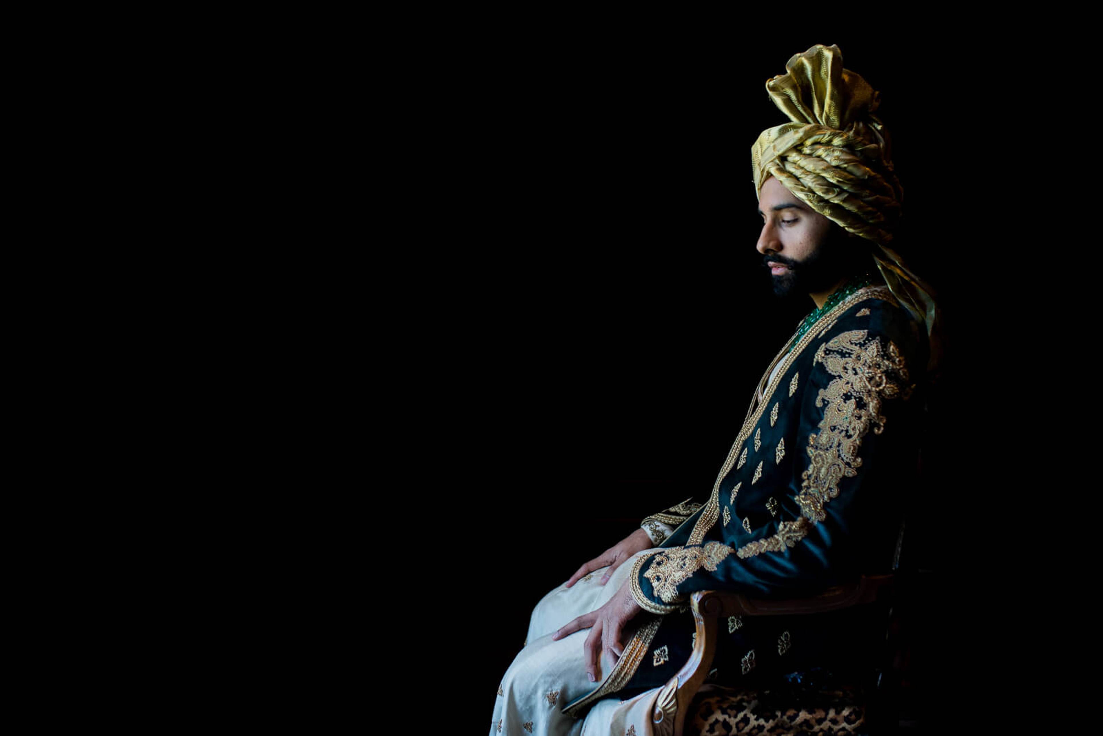 Groom in gold turban and ornate navy sherwani with embroidered details, seated in profile against black background