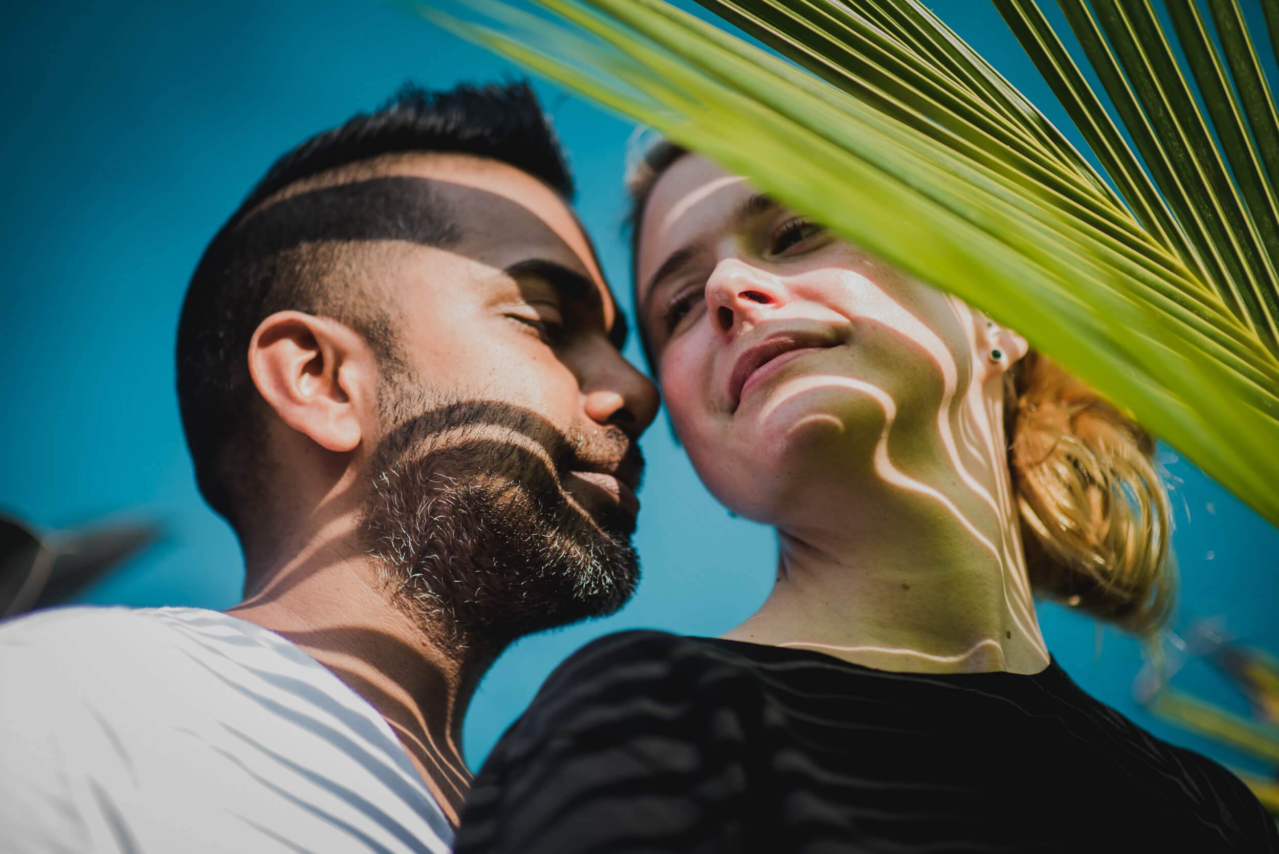 Sarah and Ravin embrace under bright sunlight beneath palm fronds in Goa, clear blue sky above