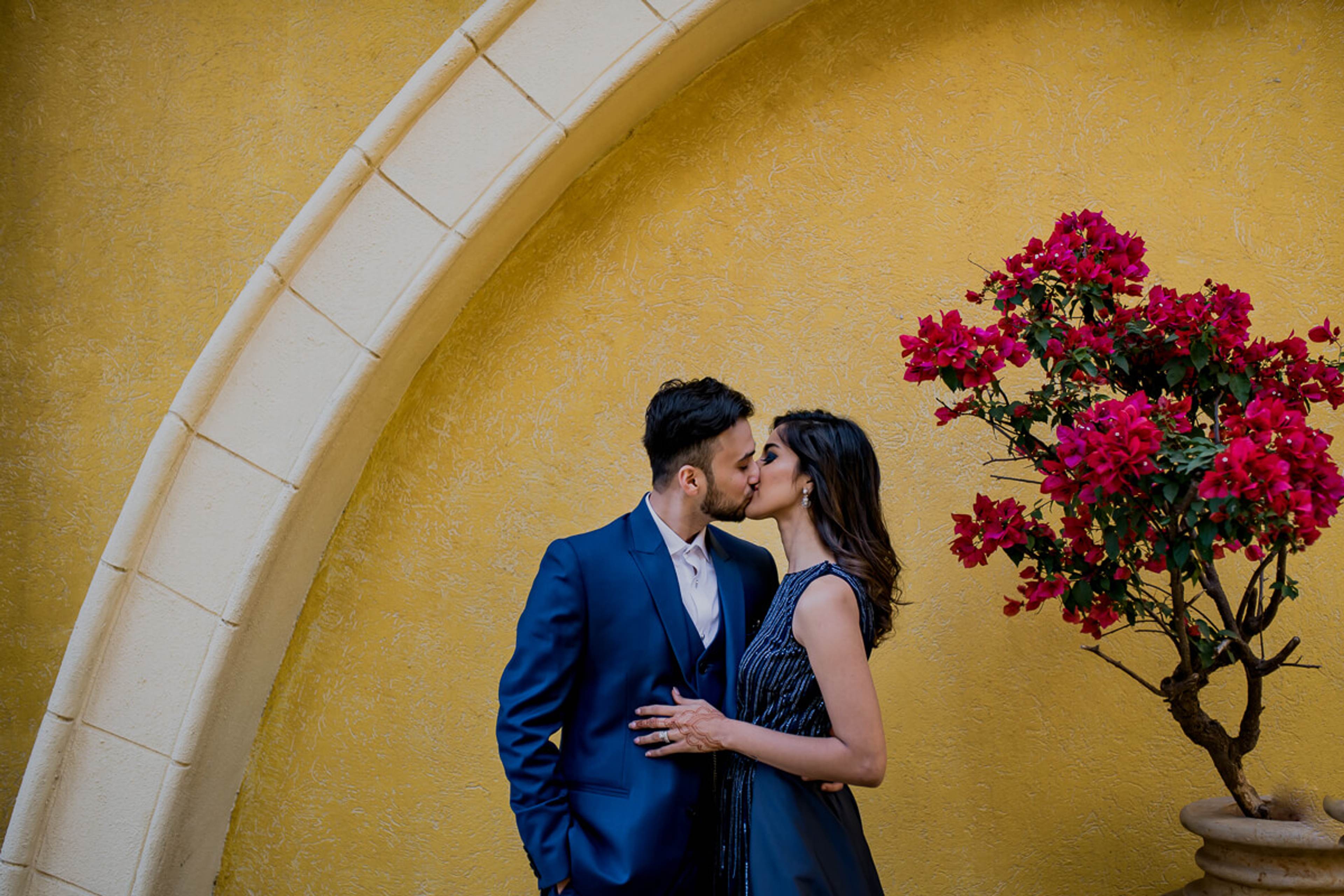 Shreya and Yash kiss beneath a curved archway, vibrant magenta bougainvillea blooming beside them against golden yellow walls