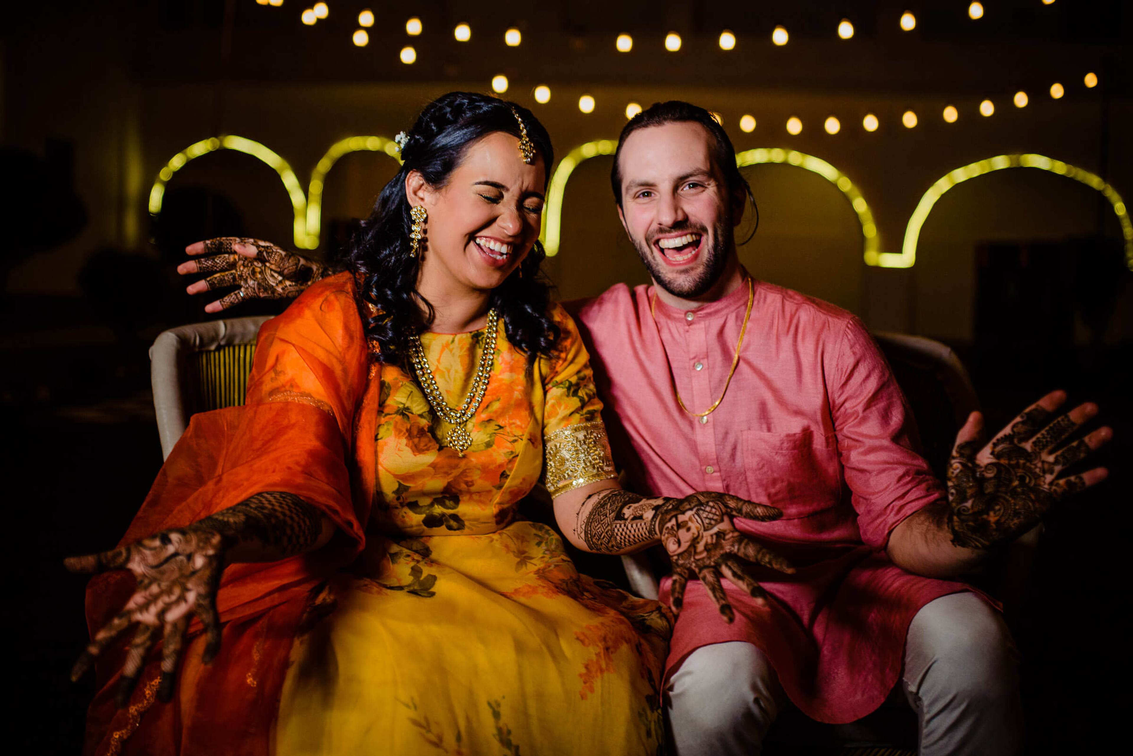 Maitrayee and Eric laugh together in traditional wedding attire with henna, warm string lights and neon arch behind them in Rajasthan