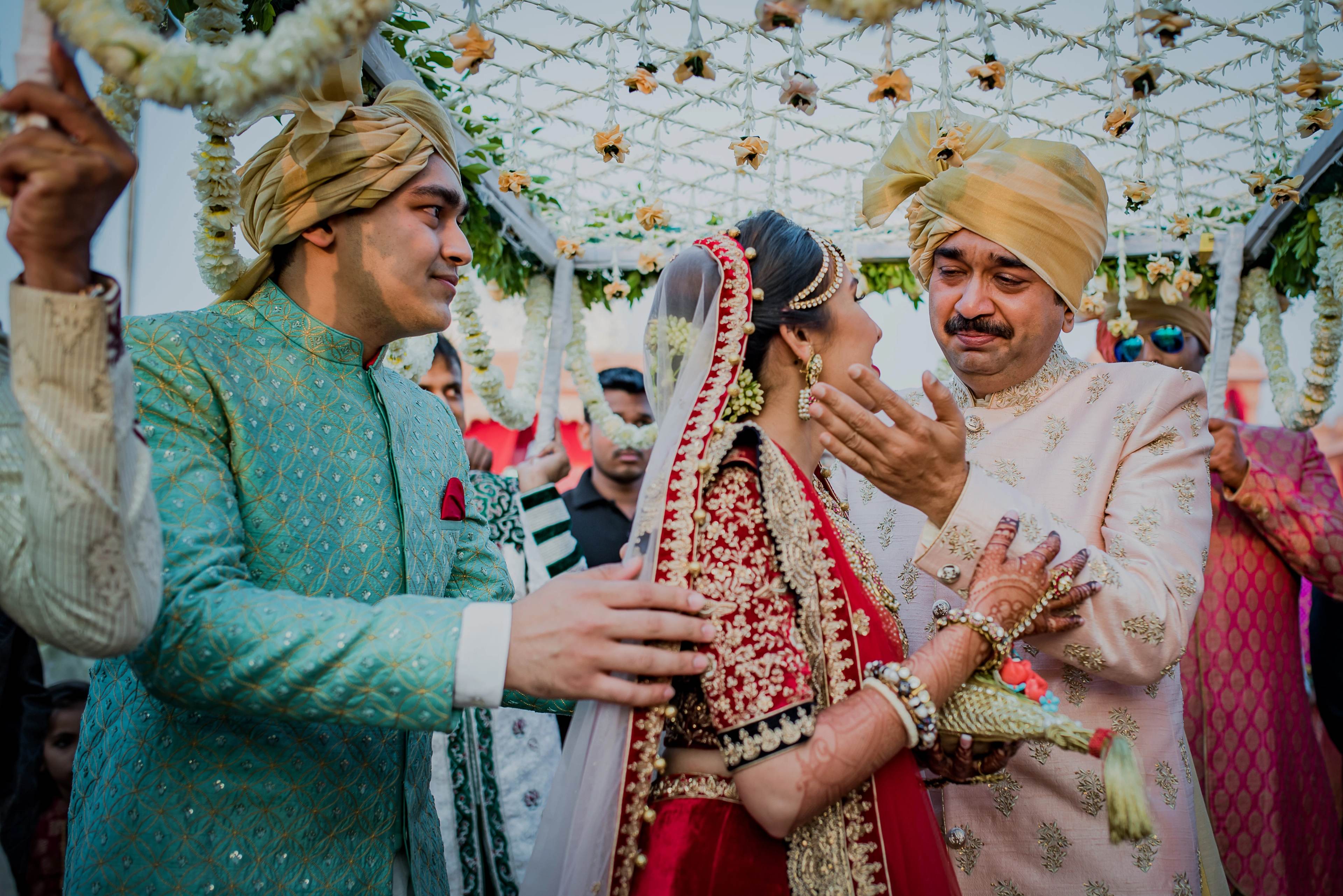 Groom in turquoise sherwani and groom in cream sherwani with bride in red lehenga under floral ceiling