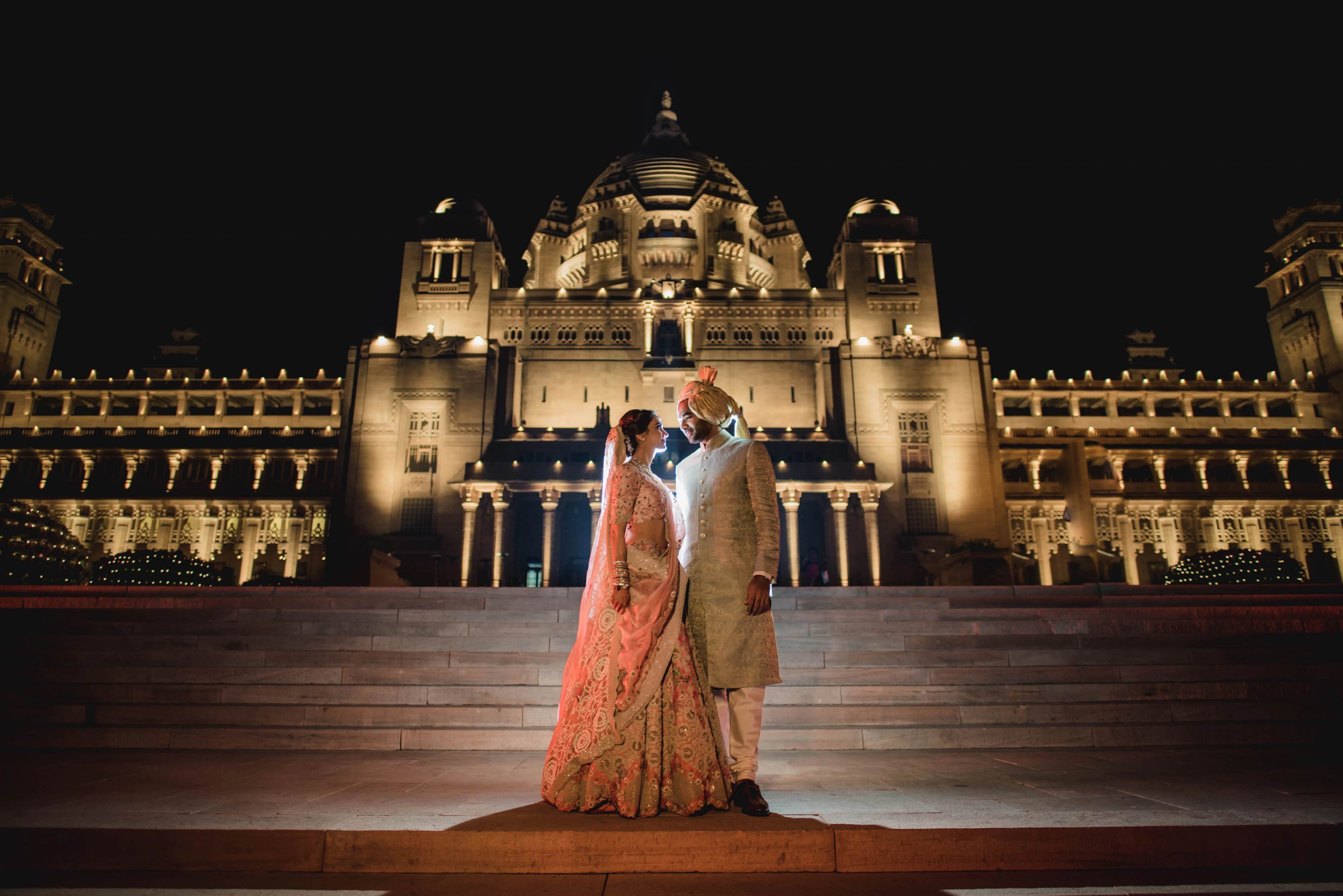 The couple stands on illuminated steps before a grand domed cathedral at night, bride in coral lehenga, groom in gold sherwani