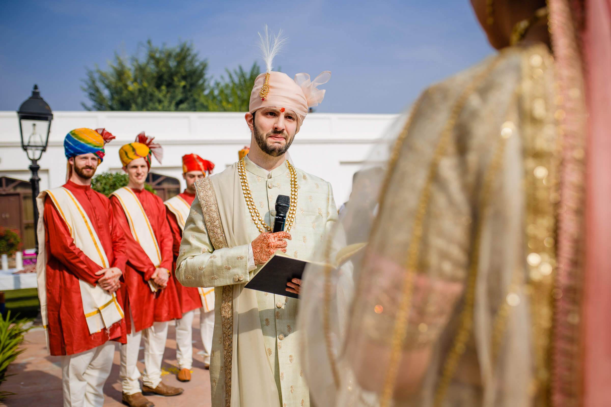 Groom in cream sherwani and turban holds tablet during ceremony in Rajasthan, groomsmen in red attire behind