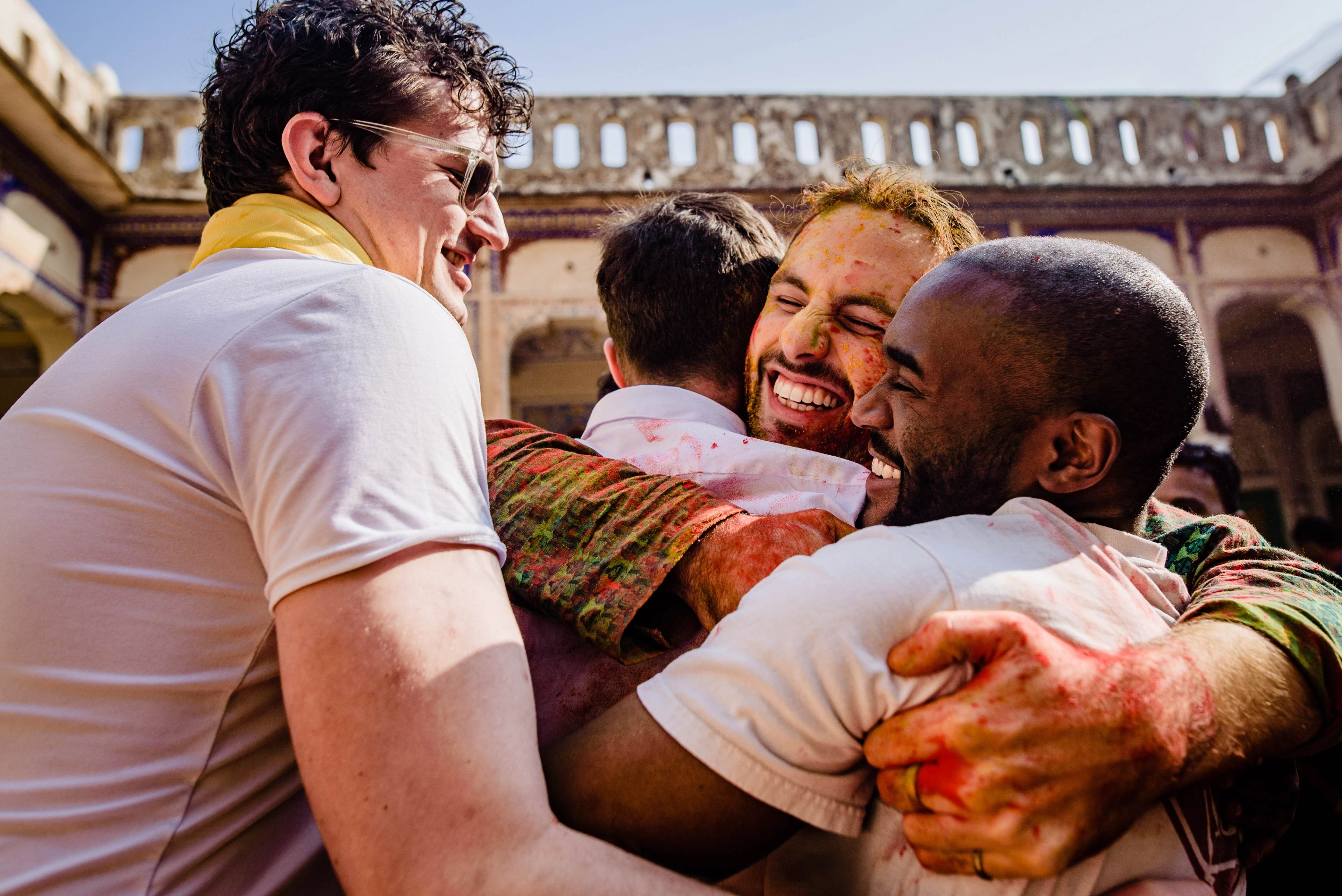 Four men embracing and laughing in sunlit courtyard with historic arched architecture