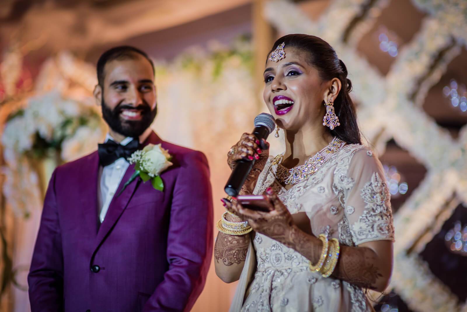 Barkha speaking into microphone in beaded white dress, Ankit smiling in purple suit at New York wedding reception