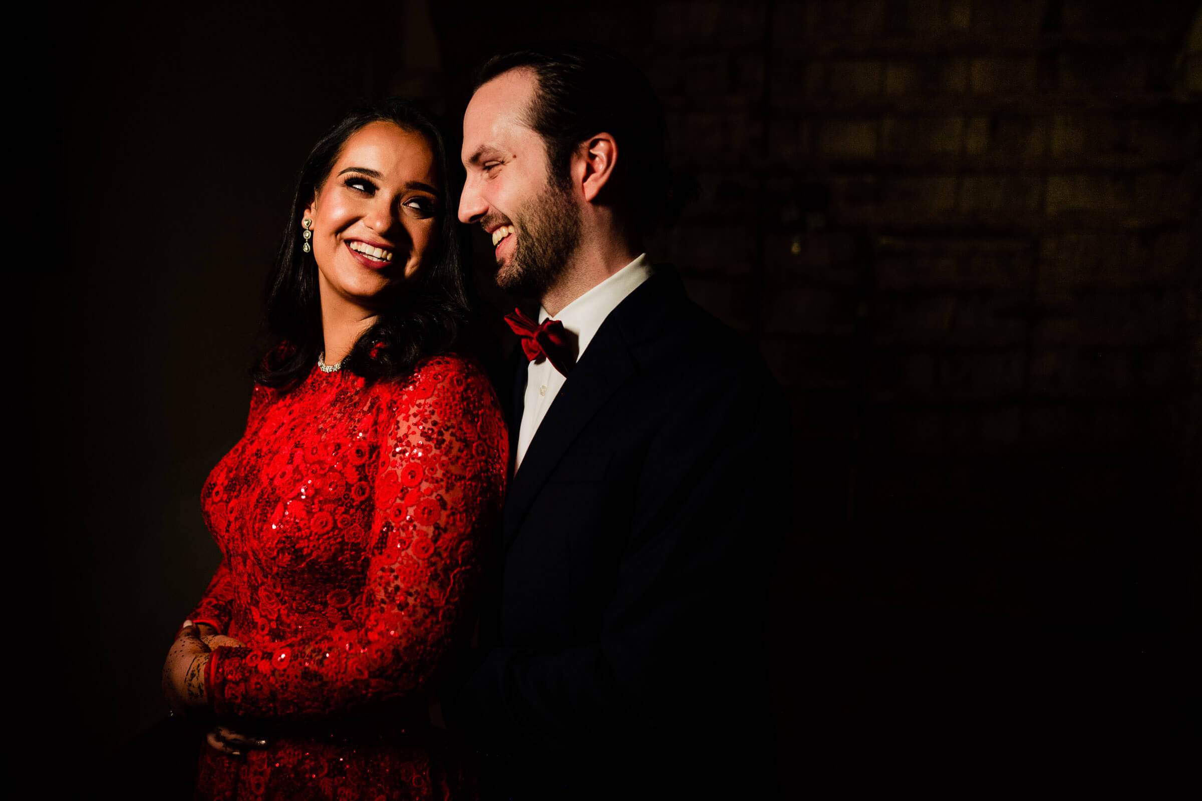 Maitrayee in a vibrant red sequined gown and Eric in a tuxedo embrace against a dark backdrop