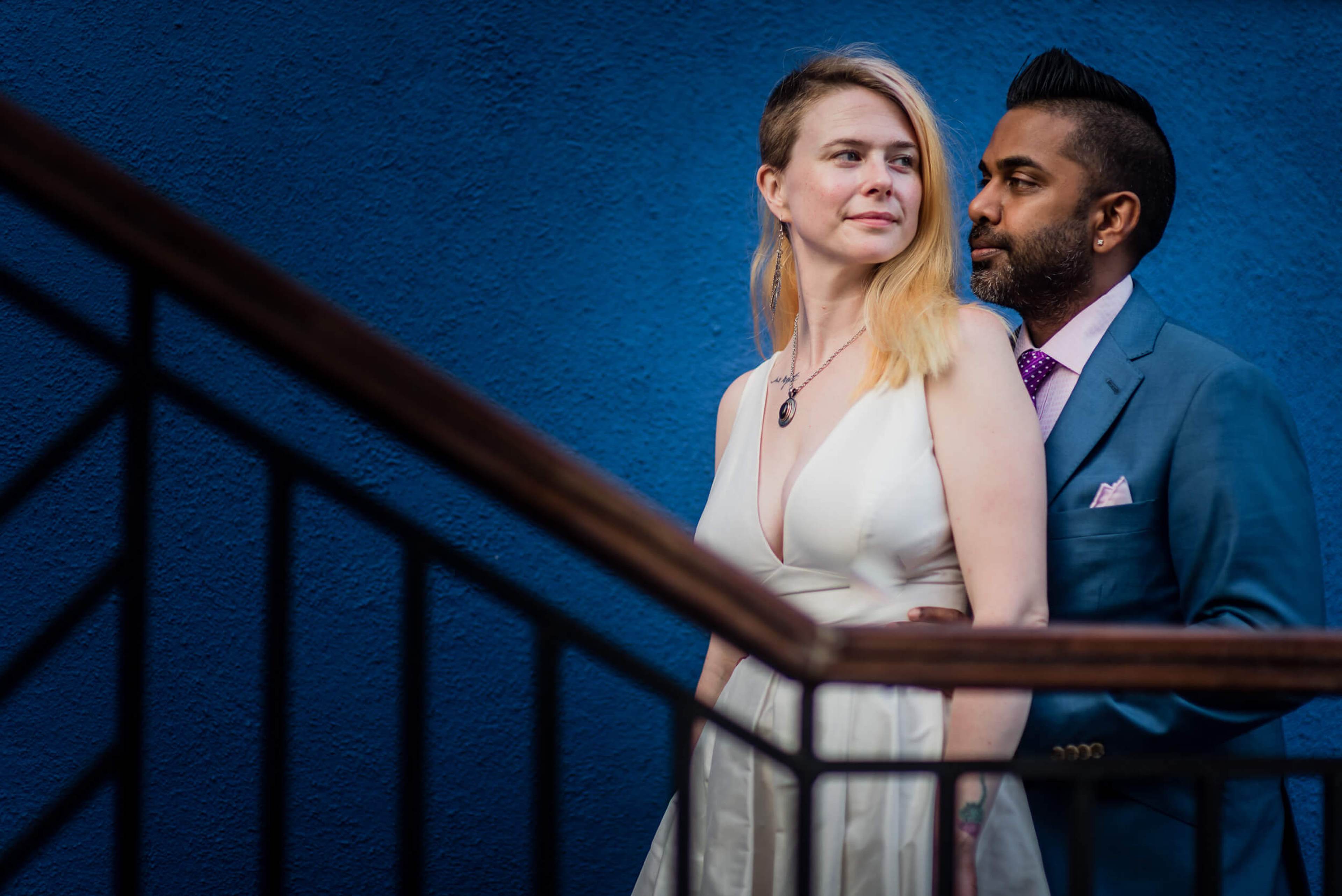 Sarah and Ravin stand together against a vivid blue wall, she in cream, he in a blue suit, with a railing in foreground